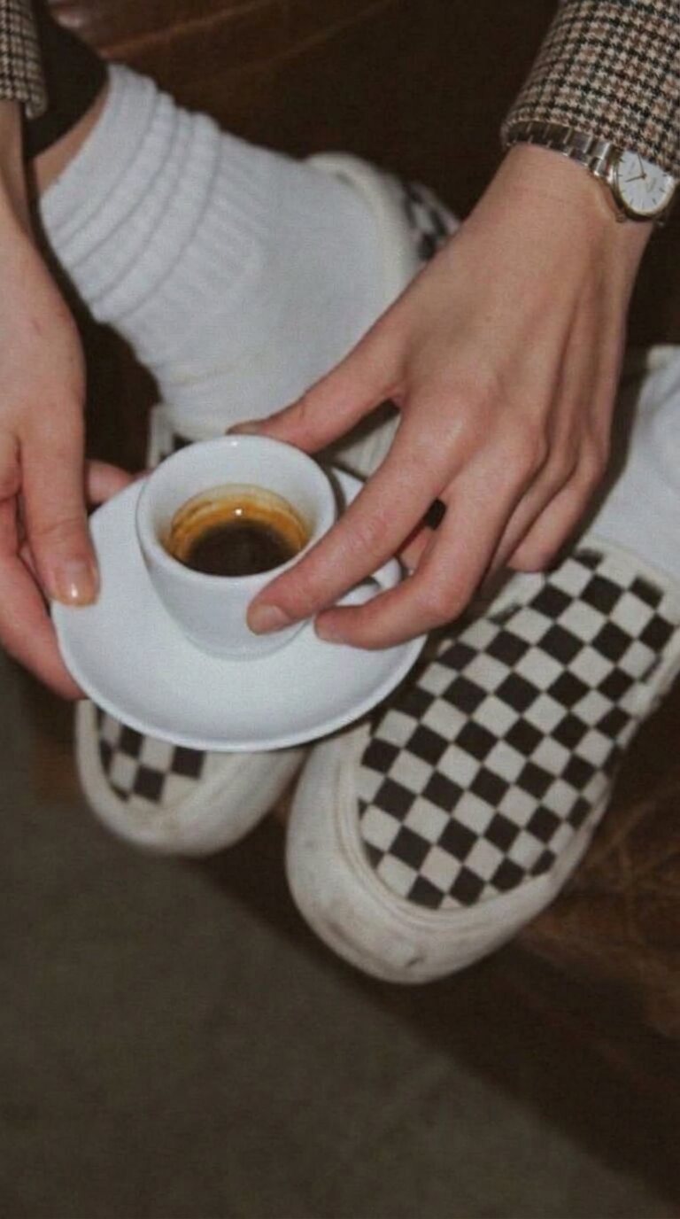 A top-down view of hands holding a small espresso cup, checkered slip-on sneakers on the floor below